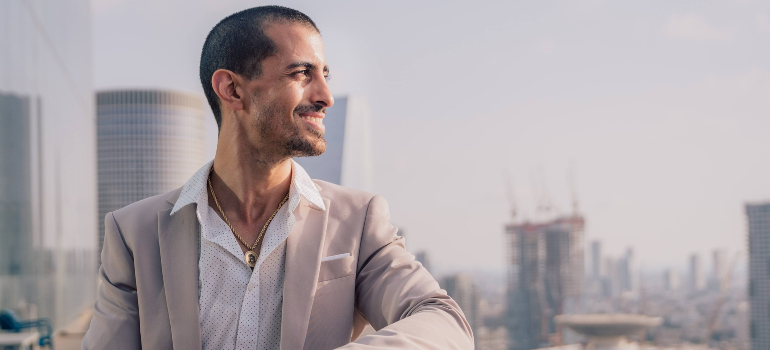 A confident man in a light suit smiles as he gazes into the distance, with a cityscape backdrop, embodying ambition and success.